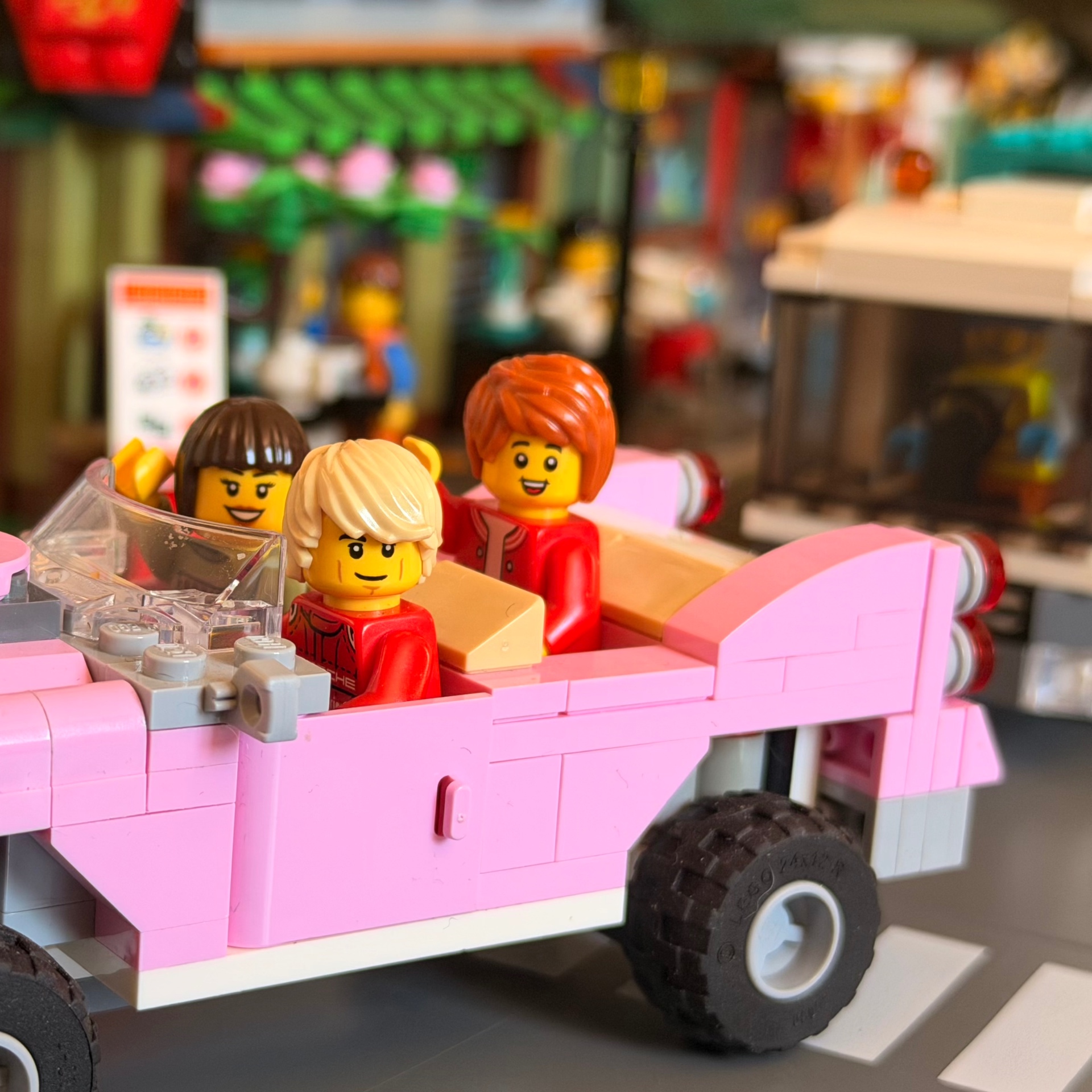 Three smiling Lego minifigures riding in a pink convertible through a colorful Lego city street